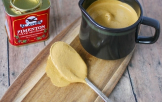A small brown pitcher of cashew cream sitting on a wooden board next to a spoon full of cream sauce.