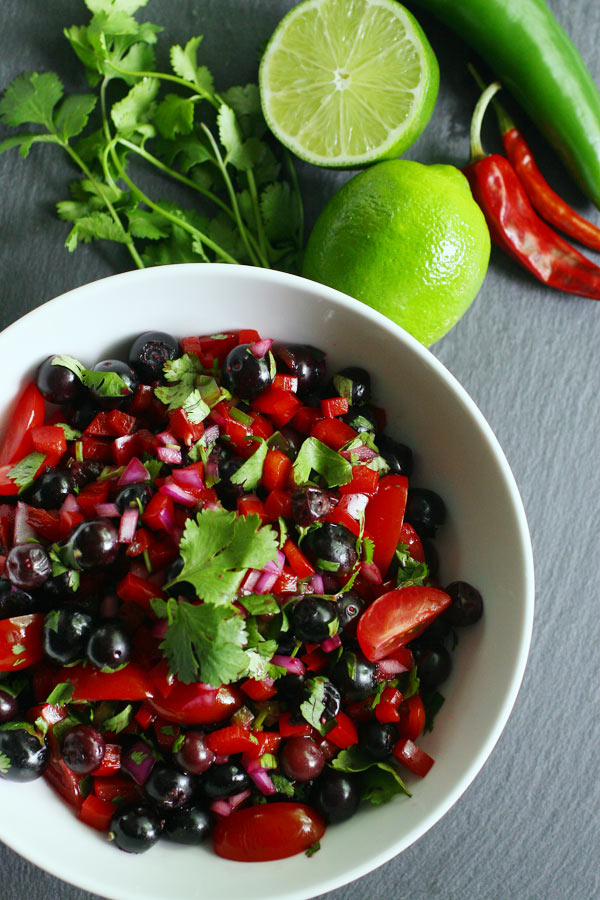 A bowl of salsa in a white bowl with limes and peppers in the background.