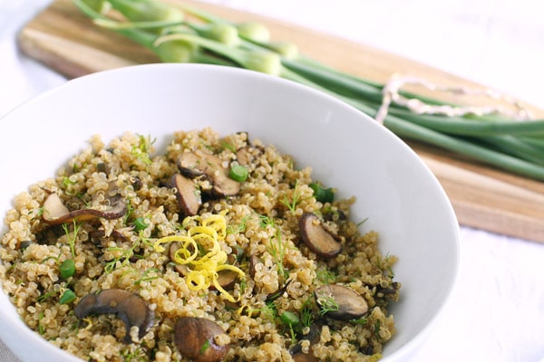 A bowl of garlic scape quinoa with a wooden cutting board with a bunch of garlic scapes sitting on it sitting in the background.