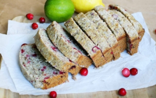 A loaf of quick bread sitting on parchment paper on top of a wooden table with lemons, limes, and cranberries strewn about.