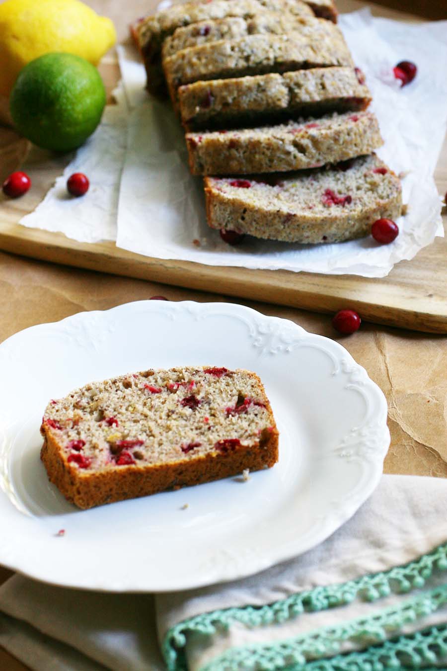 A loaf of quick bread sitting on parchment paper on top of a wooden table with lemons, limes, and cranberries strewn about.