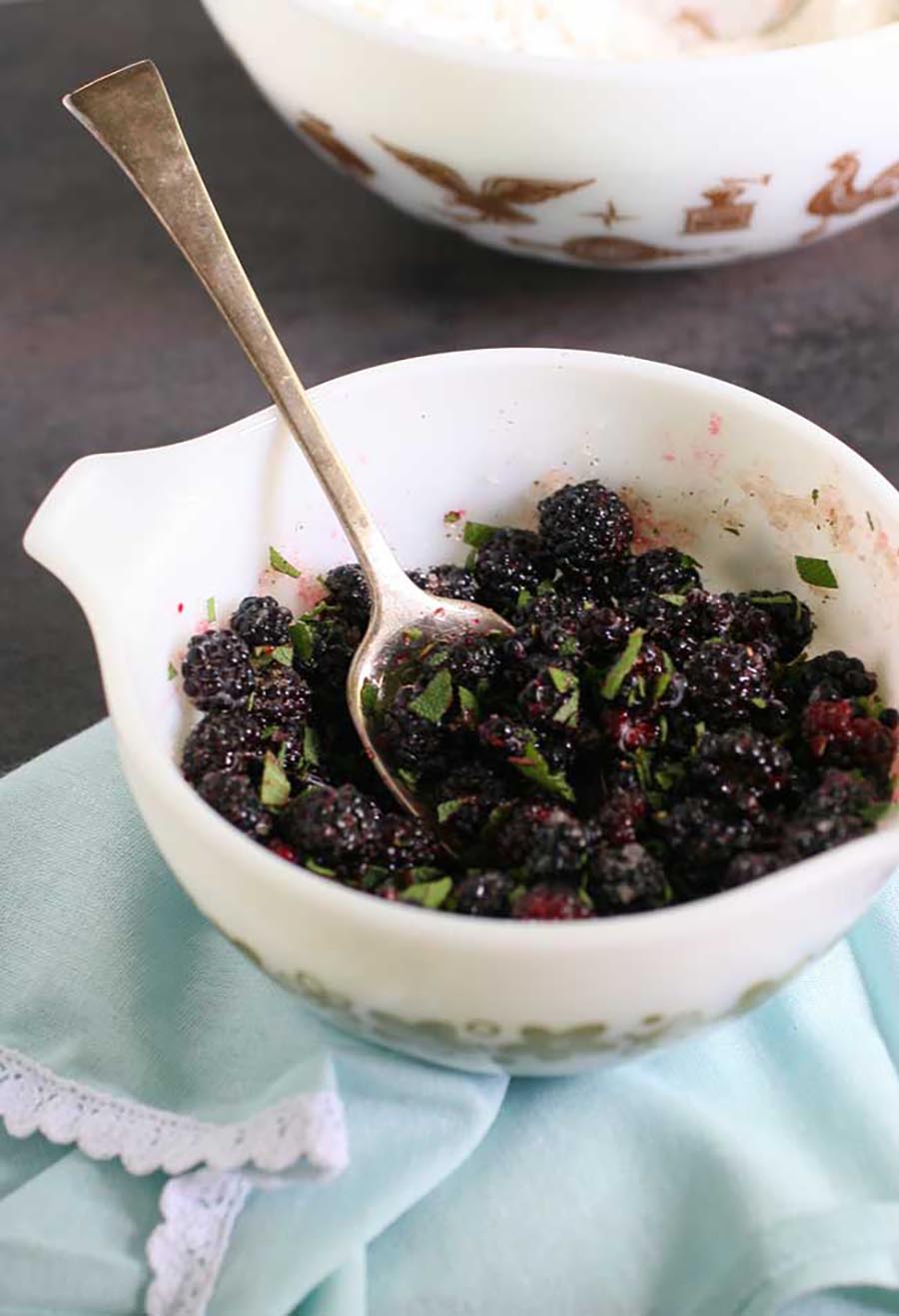 A white bowl of blackberries sitting on a blue tea towel with a white bowl of flour in the background.