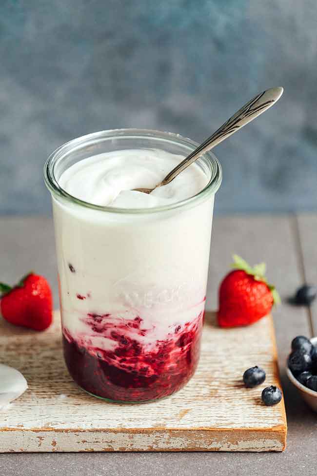 A glass of yogurt sitting on a wooden plank on a wooden table with berries in the background.
