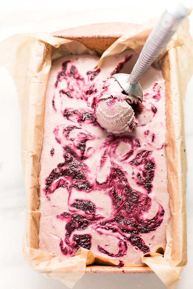 An overhead picture of a rectangle pan with bright pink vegan ice cream with swirls of red fruits throughout and an ice cream scoop sitting on one side.