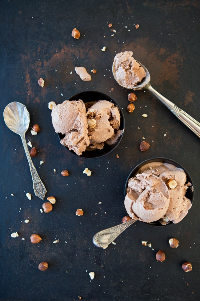 An overhead picture of a bowl of ice cream with several spoons full of ice cream and nuts scattered about sitting on top of a black background.