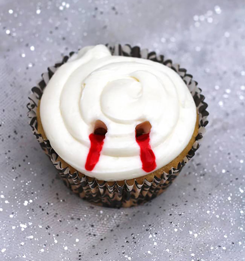 A closeup of a cupcake with white frosting and two "bites" with red oozing from them sitting on a grey speckled countertop.
