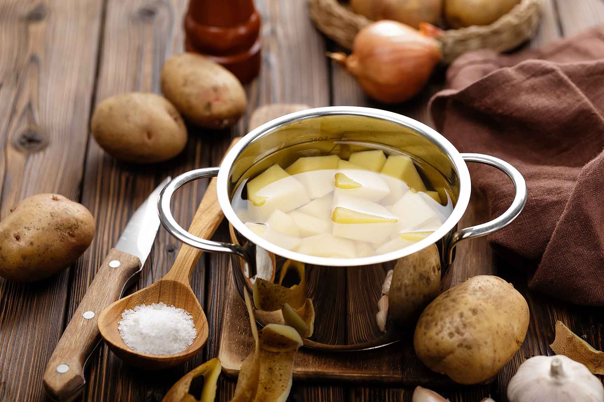 A pot full of water and cut potatoes sitting on a wooden table surrounded by whole potatoes, onions, garlic, and a knife.