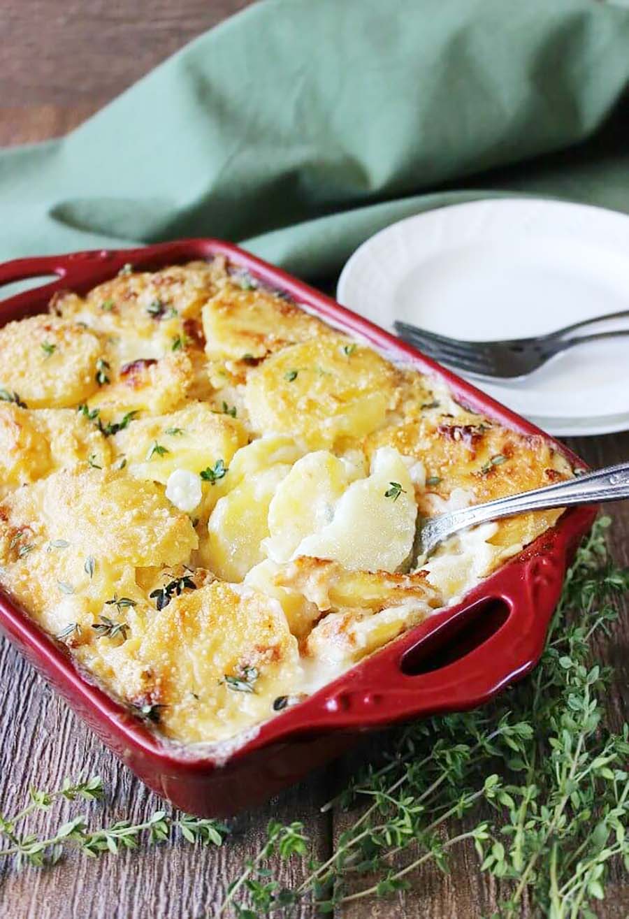Scalloped potatoes in a red casserole dish sitting on a wooden table with a stack of plates, cutlery, and napkins nearby.