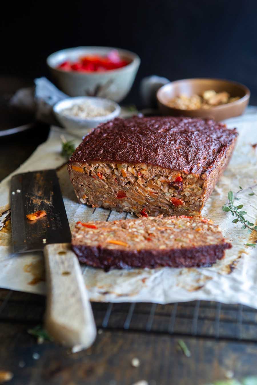 A cutting board sitting on a wooden table with a piece of parchment paper wrapped lentil loaf sitting on top with a knife and condiments sitting in the background.