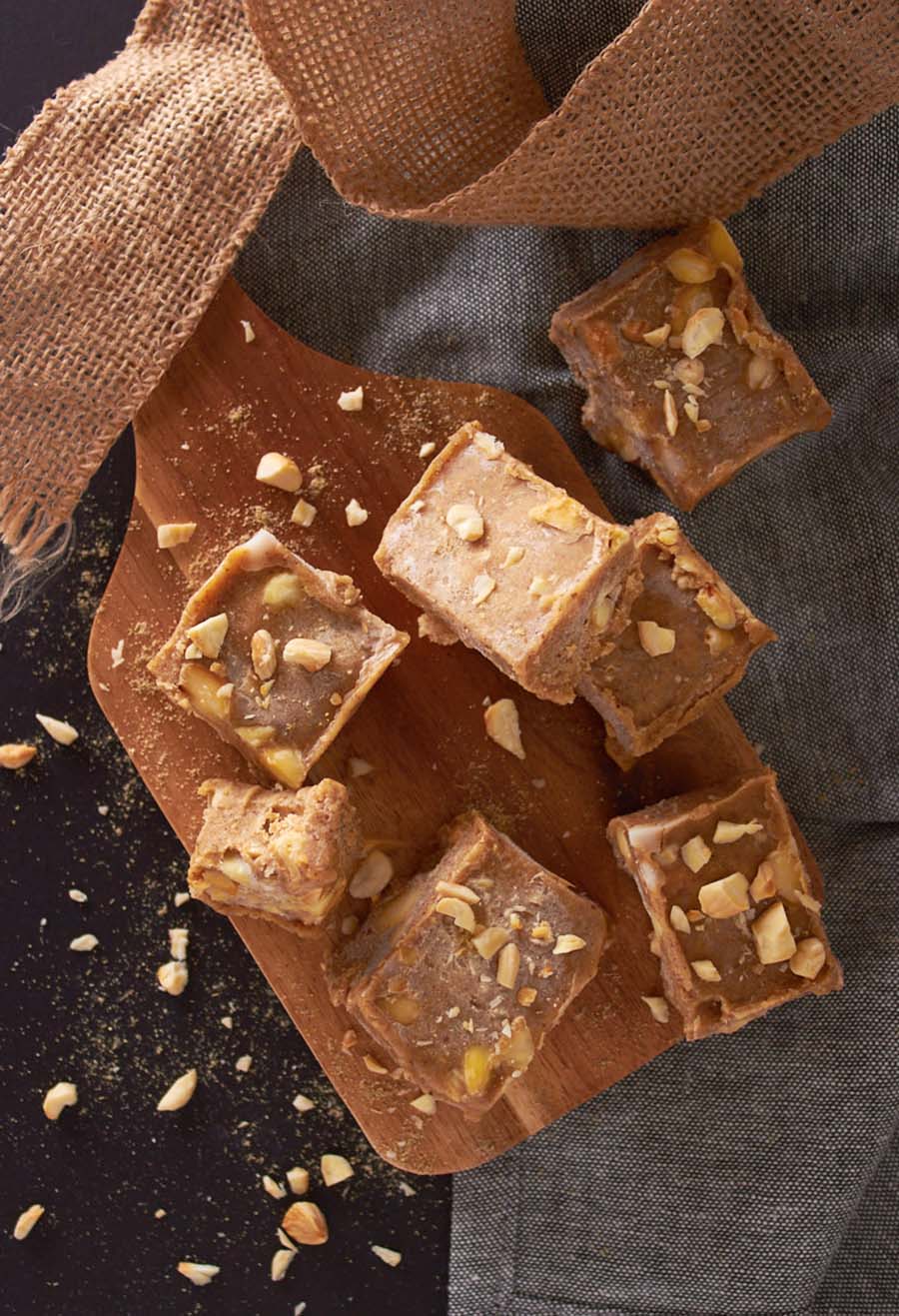 Blocks of fudge sitting on a wooden cutting board that is on top of a black counter with nut pieces strewn about.