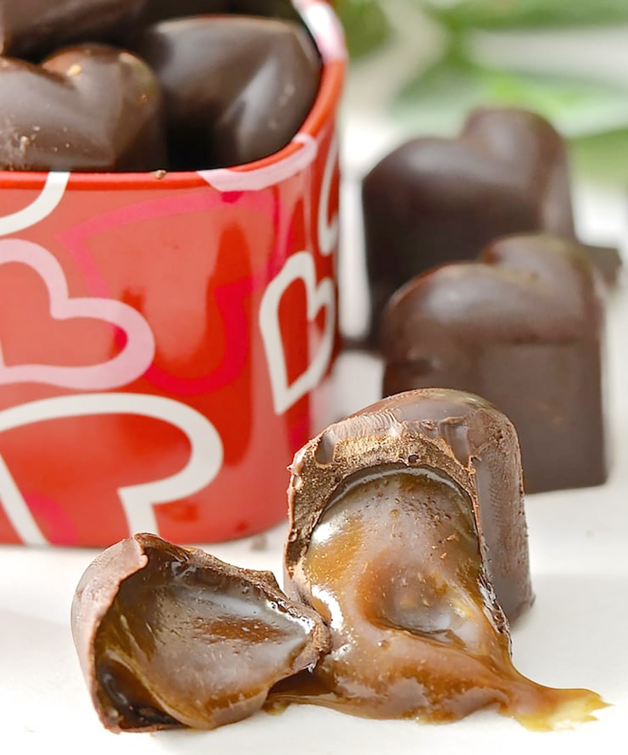 A close-up of a chocolate caramel that has been broken in two with the caramel oozing from it with a tin of chocolates in the background.