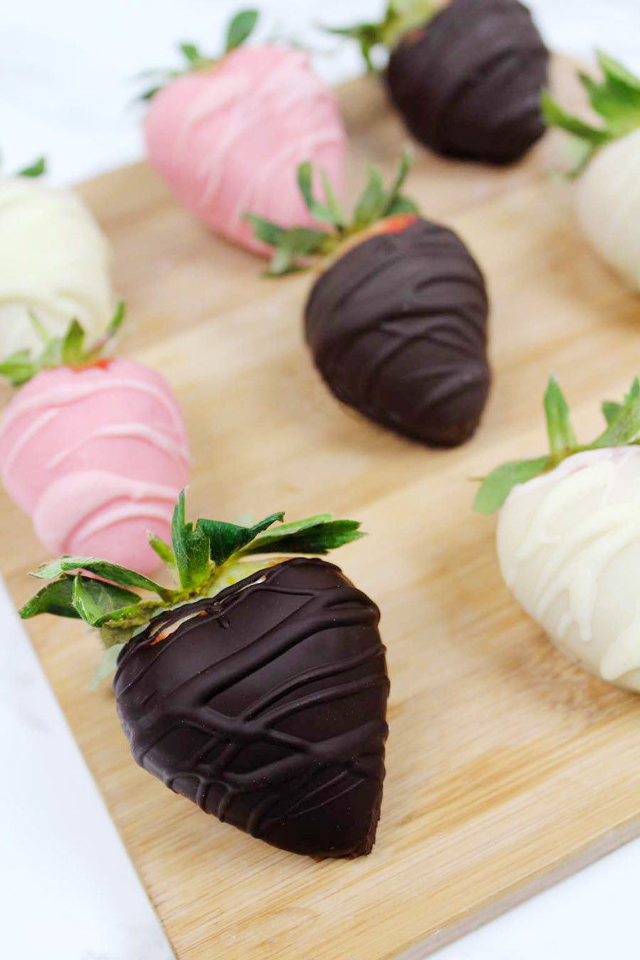 A group of chocolate covered strawberries arranged on a wooden cutting board sitting on a white countertop.