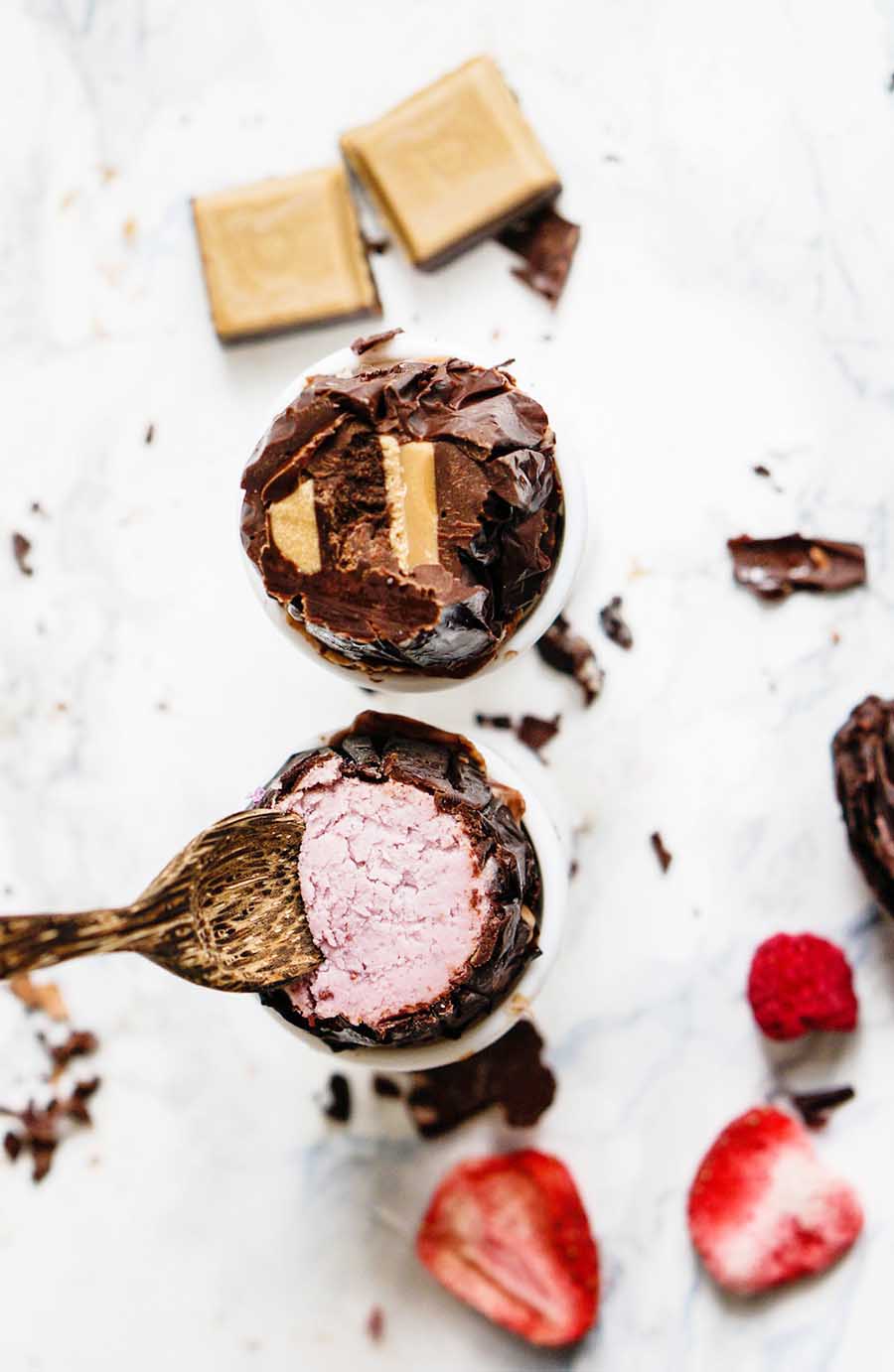 Overhead view of a spoon scooping out a pink dessert from inside a half of a chocolate egg with various chocolate pieces and fruit pieces laying around.