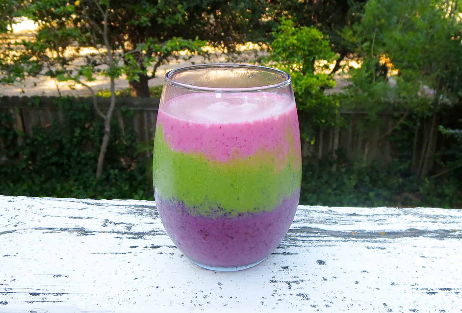 A colorful smoothie in a clear glass sitting on a white outdoor railing.