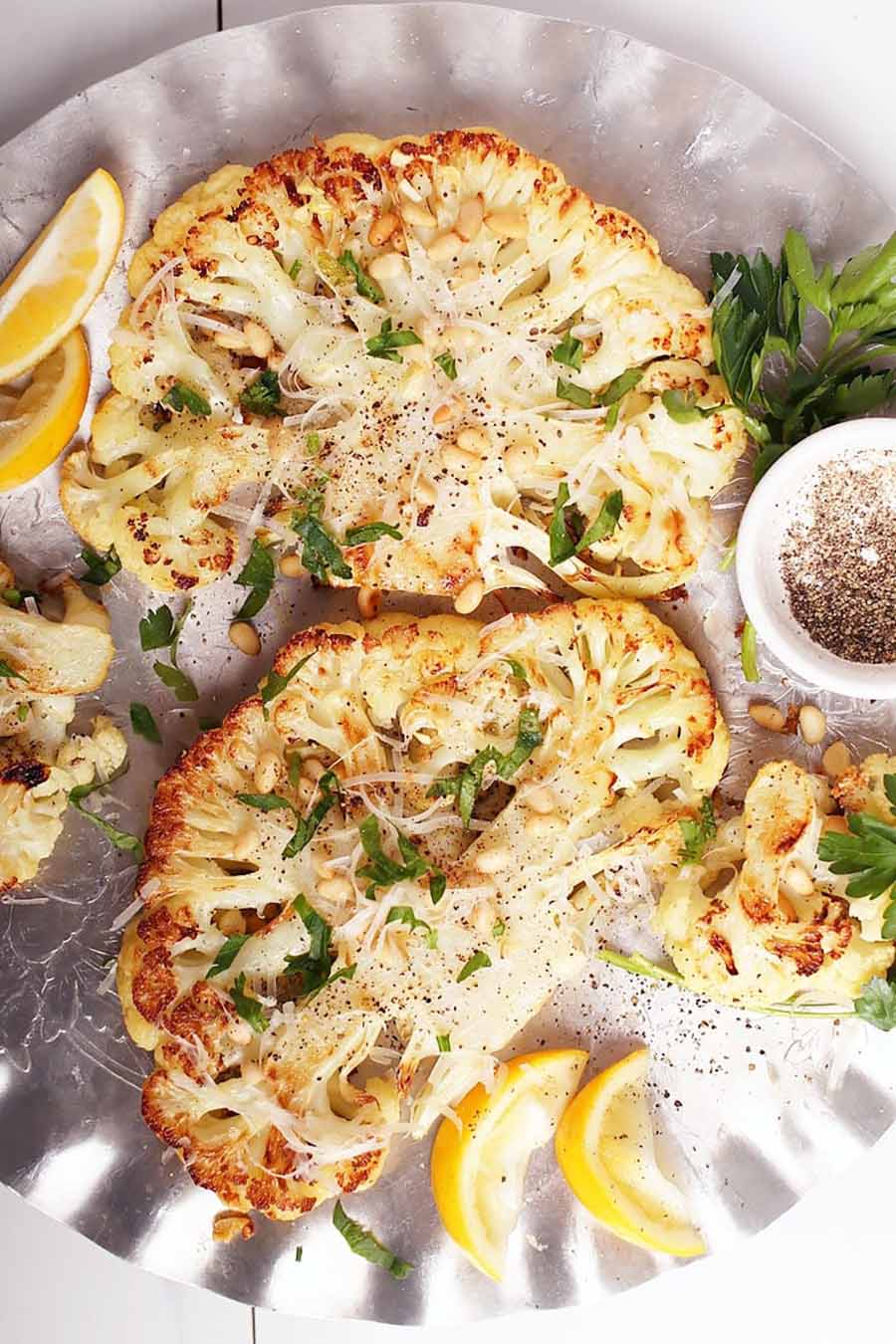 Overhead view of cauliflower steaks sitting on a silver platter with a few lemon wedges nearby.