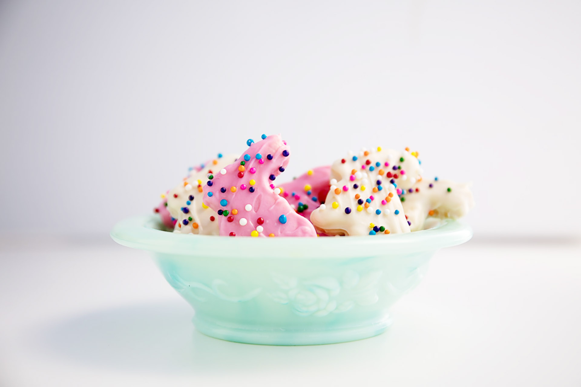 A light green glass bowl filled with pink and white frosted animal cookies with colorful sprinkles on top.