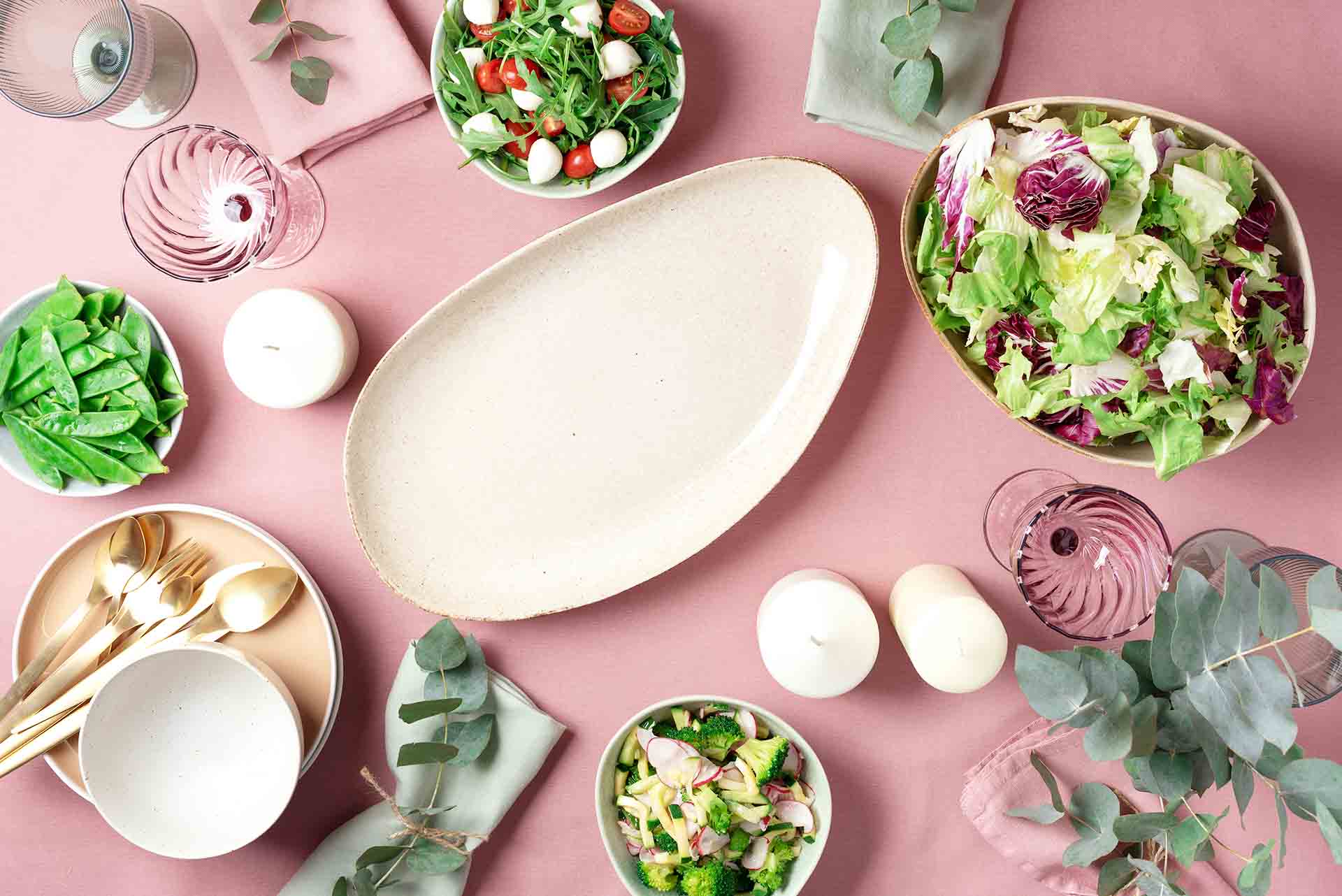 Serving plates, bowls with salads and vegetables dishes, eucalyptus flowers on pink shabby background.