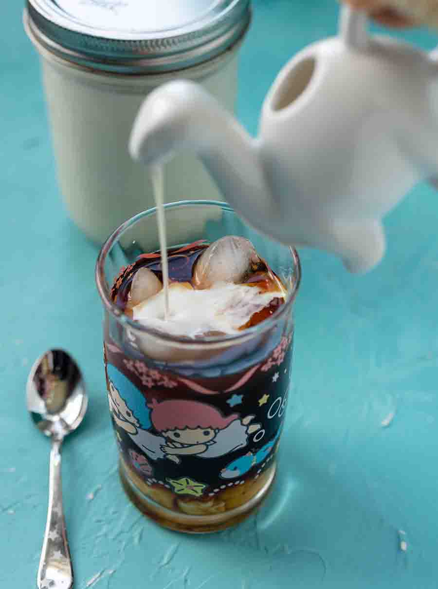 A white dinosaur-shaped creamer pouring into a glass cup filled with coffee