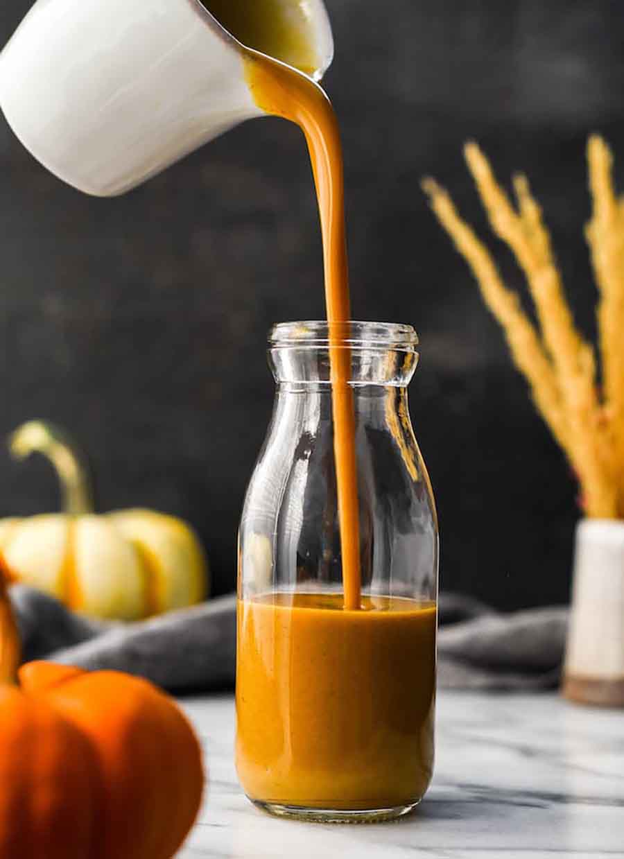 A carafe of creamer being poured into a glass jar on a table with pumpkins