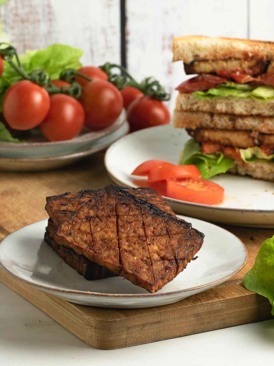 A plate of tempeh bacon sitting on a table with fresh ingredients and a sandwich